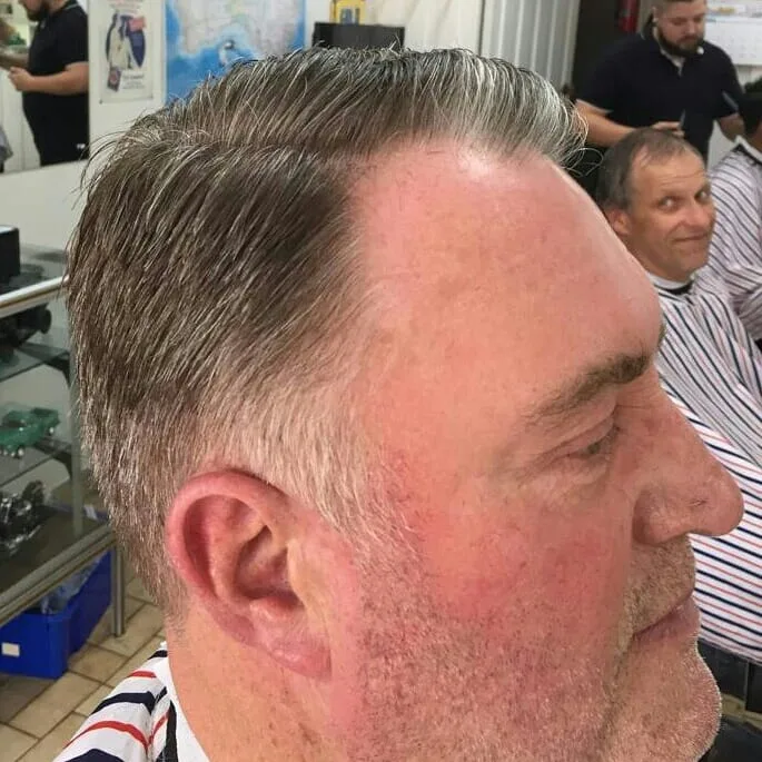 Older man's grey slicked side-profile; a second barber at work in the background