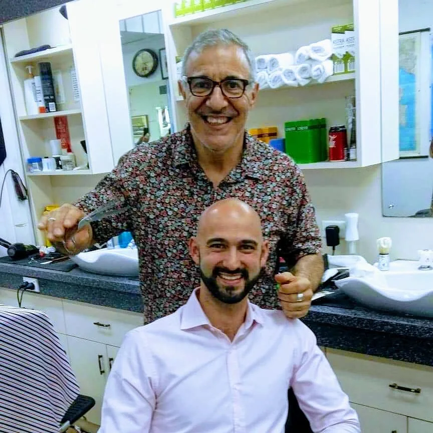 The barber at work — older man with glasses, warm smile, scissors in hand; client laughing in the chair