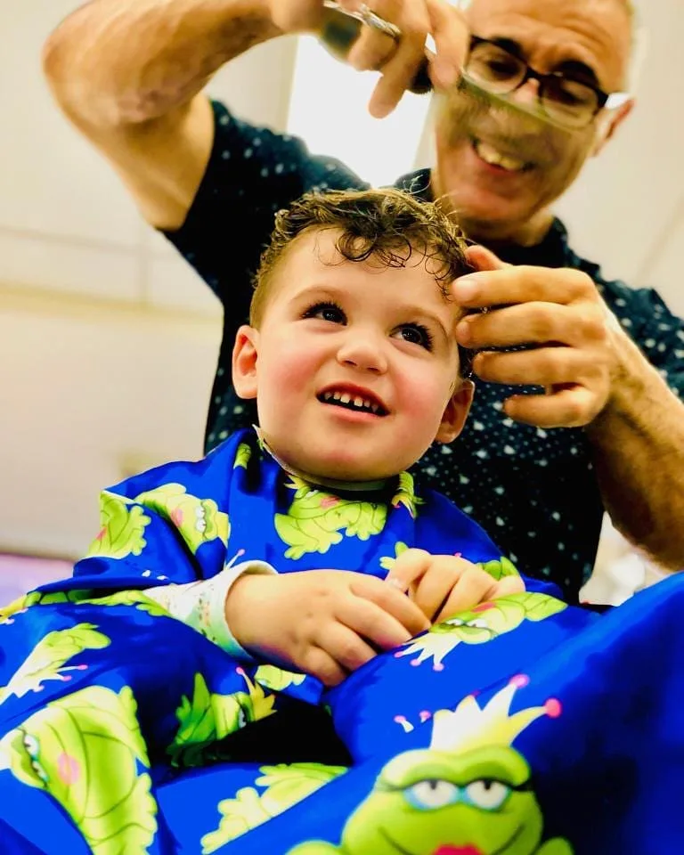 Barber cutting a toddler's curly hair — child in a blue frog cape, grinning up